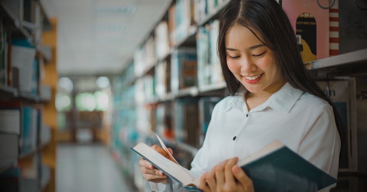 Young woman smiling while reading a book in a library aisle, symbolizing learning, career growth, and resume-building opportunities for community workshops.